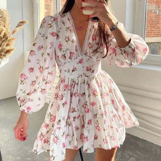 A woman wearing a white long-sleeve mini dress with a pink and green floral print, featuring a lace-trimmed V-neckline and a flared skirt, shown in a mirror selfie.